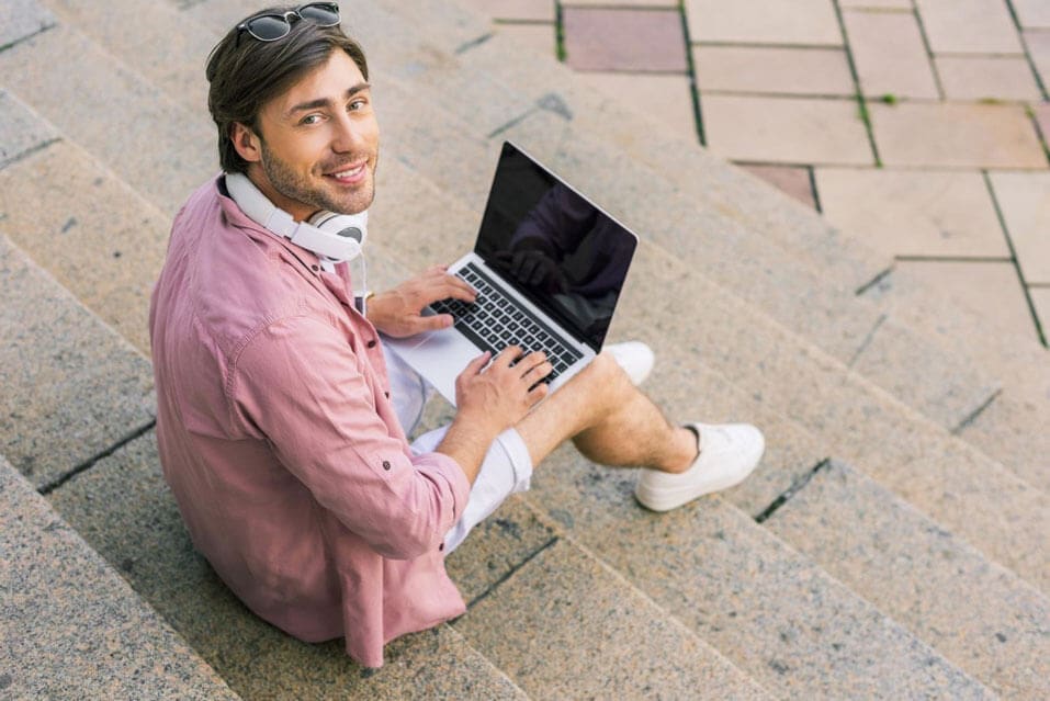 A guy is staring at the camera with headphones around his neck and his laptop open