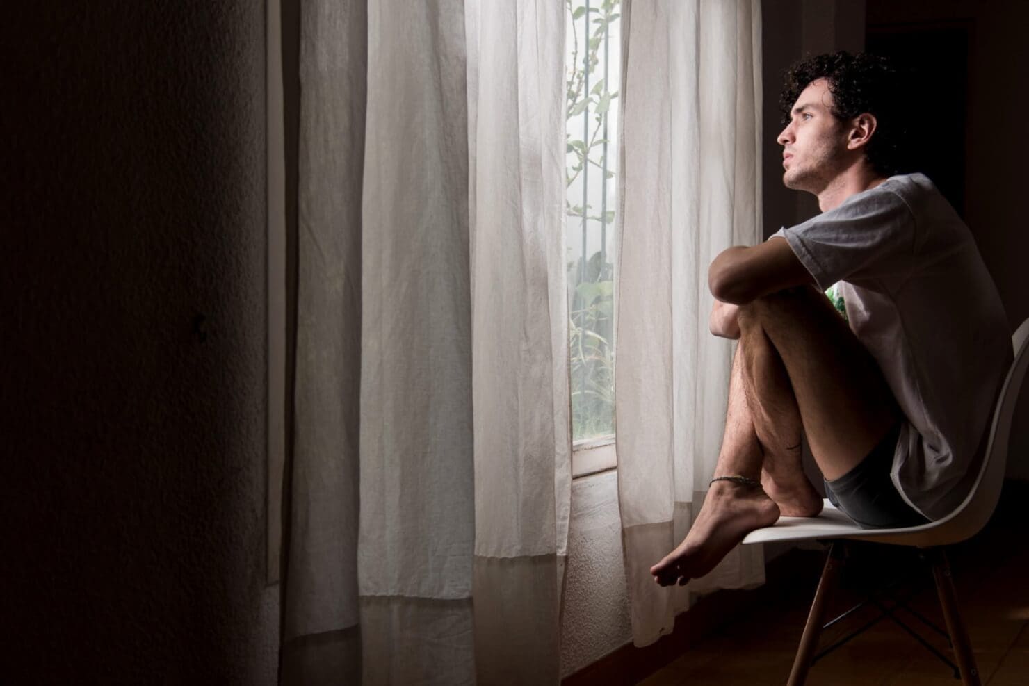 A young guy is sitting on a chair looking out his window.