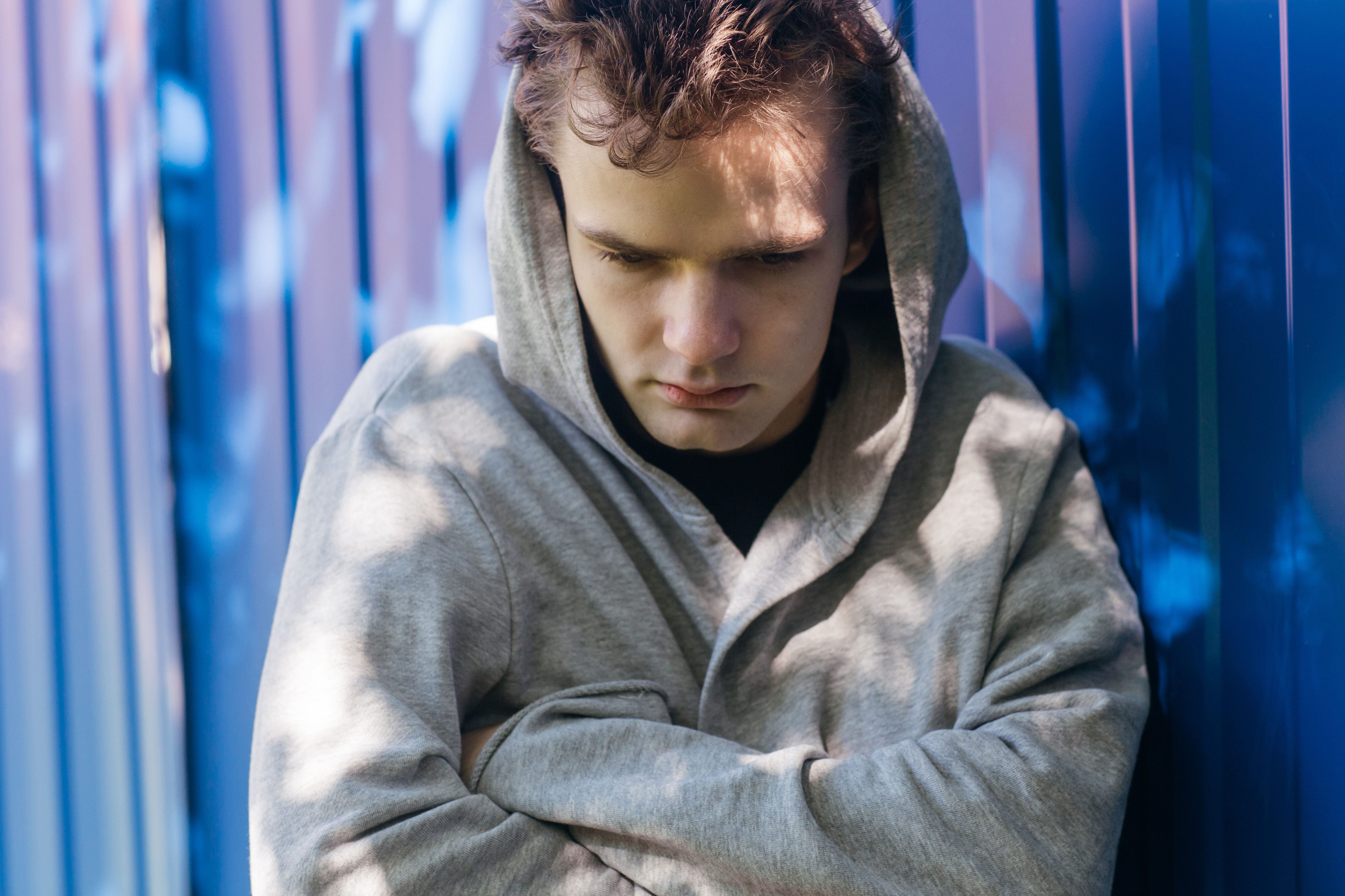 A young man in a hoodie seeking addiction recovery support leans against a blue wall, contemplating his journey towards wellness in an outpatient program.