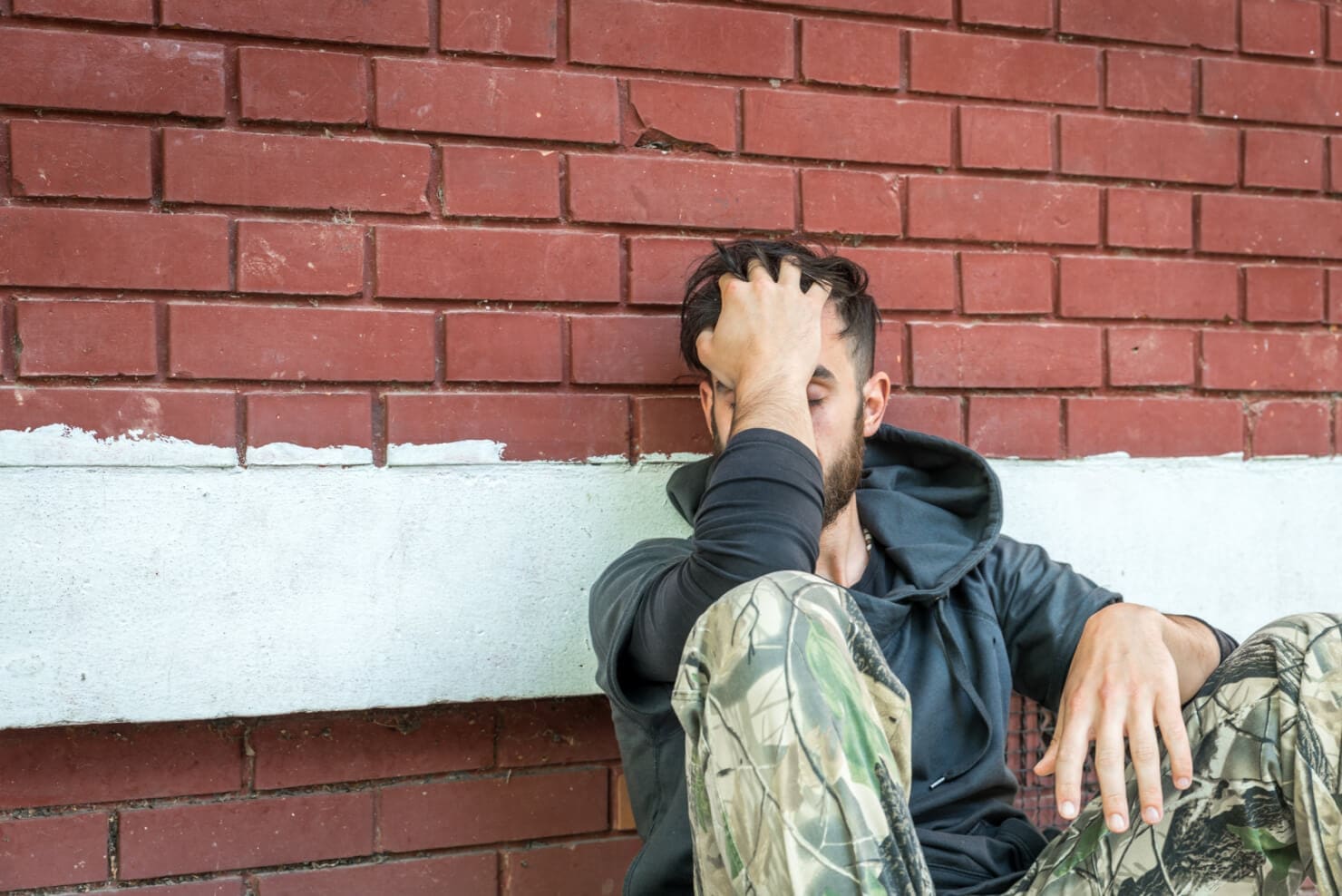 A man sitting on a brick wall at an addiction recovery center, with his hand on his face.