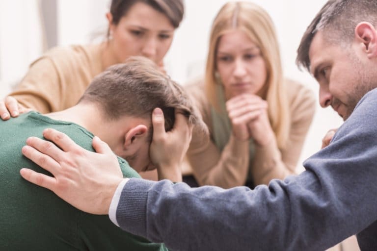 A group of people attending an addiction recovery center, sitting around a man who is being comforted.