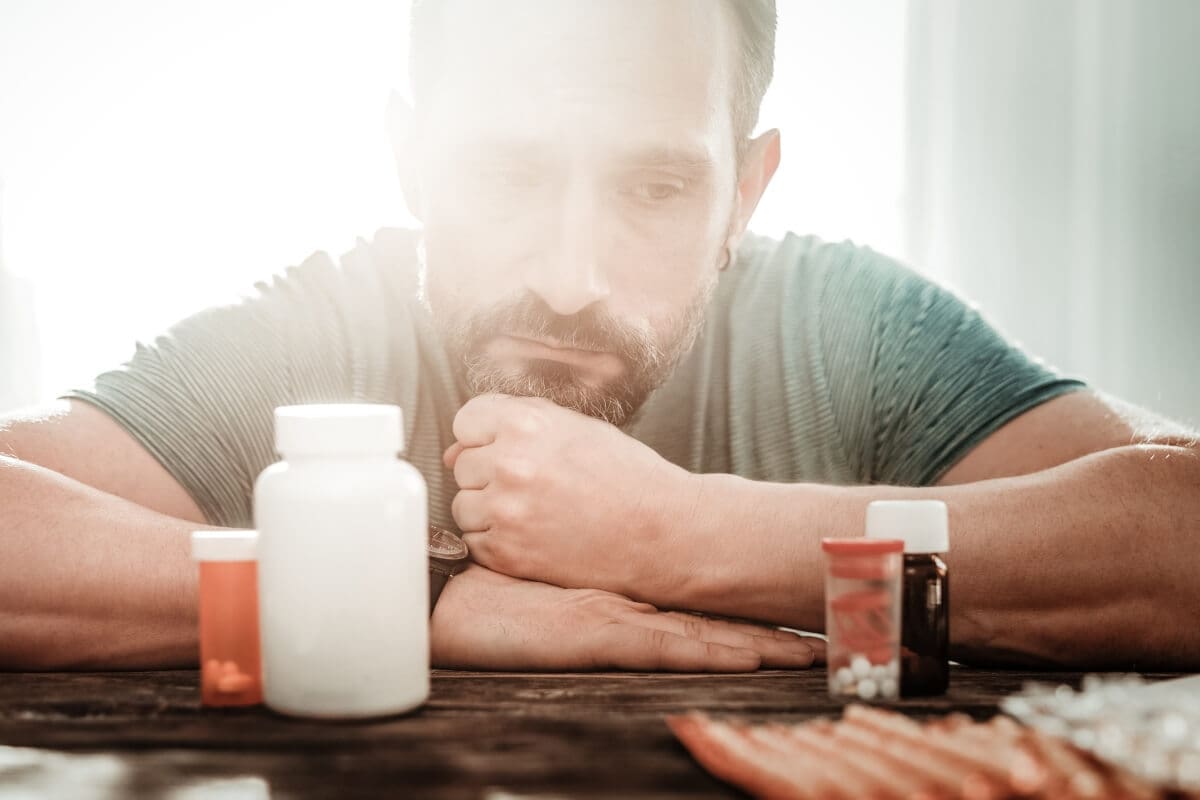 A man is sitting at a table before joining a drug detox center, surrounded by pills and medicine during his rehab journey.
