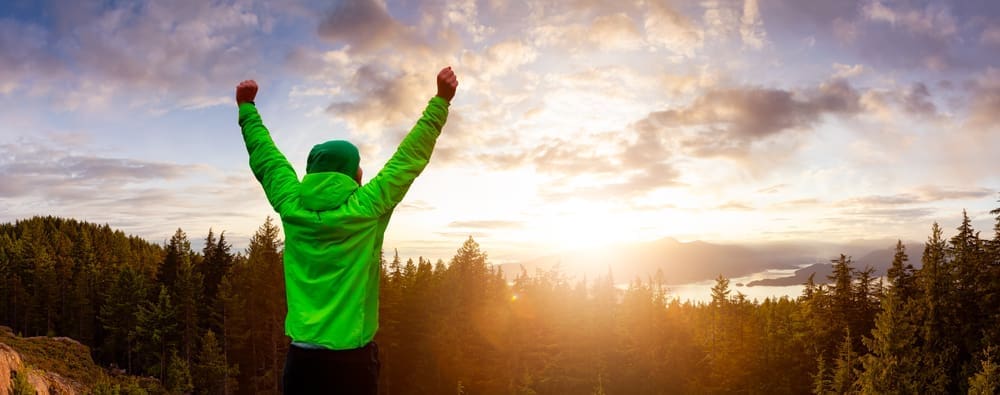 A man standing on top of a mountain with his arms raised in the air after recoving from addiction.